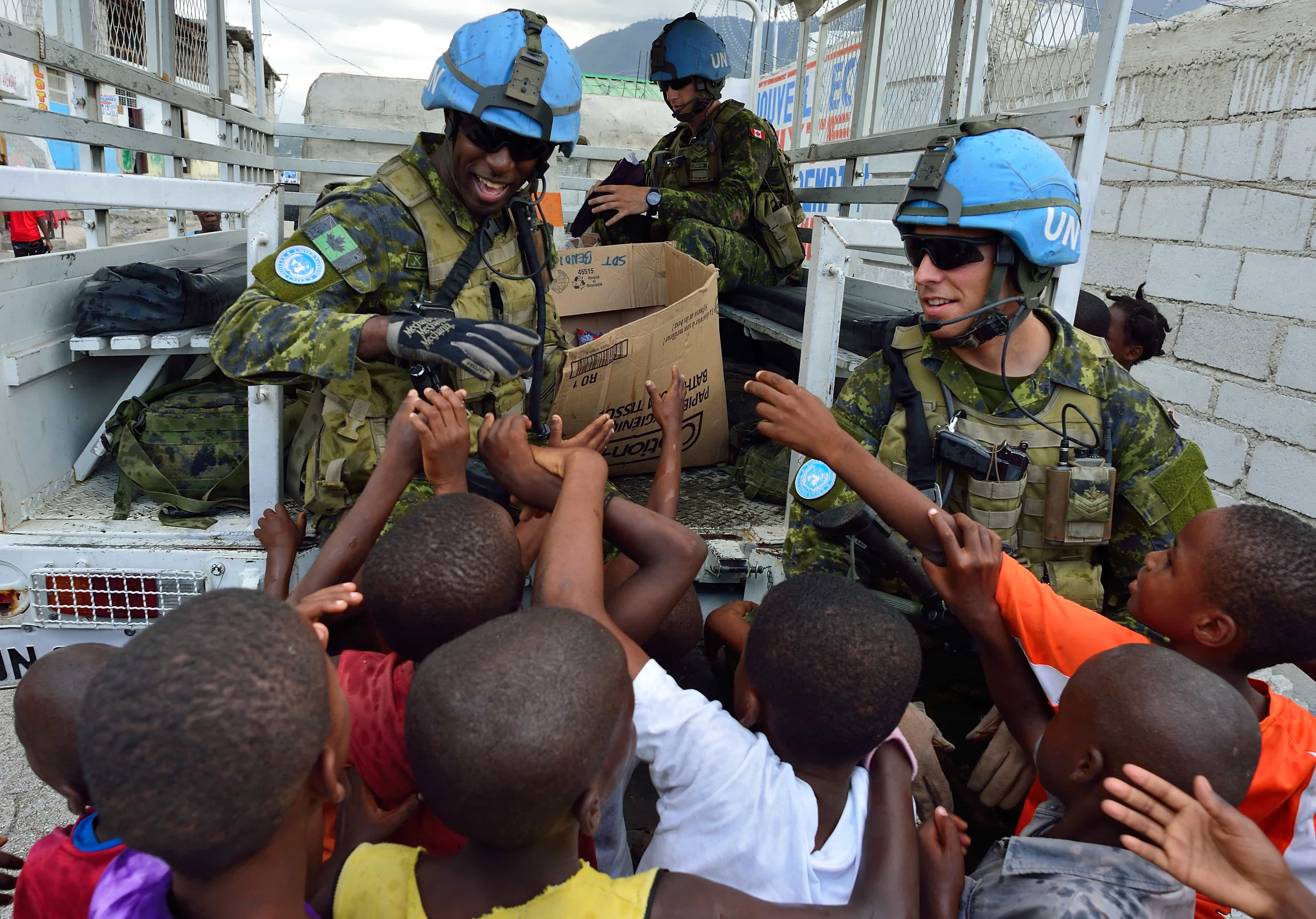 De nombreux enfants noirs entourent un homme noir souriant en uniforme (Casque bleu), sous le regard d’un homme blanc également en uniforme de maintien de la paix.